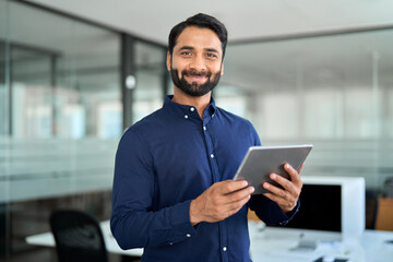 Smiling professional Indian businessman using tab computer working standing in office. Confident professional business man entrepreneur holding digital tablet tech device working, looking at camera.