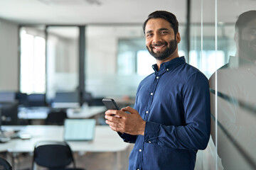 Smiling Indian businessman using mobile cell phone working standing in office. Happy professional business man making banking transaction payment on smartphone working on cellphone at work. Portrait. - obrazy, fototapety, plakaty