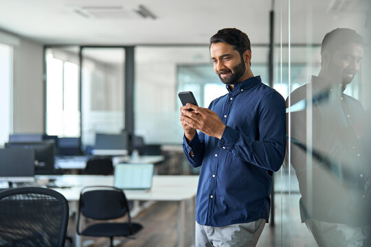 Busy Indian businessman using mobile cell phone working standing in office. Smiling professional business man making banking transaction payment on smartphone working on cellphone at work.