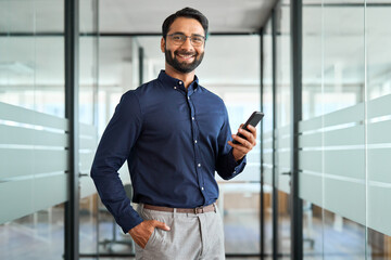 Happy Indian business man holding mobile cell phone device looking at camera standing at work. Smiling businessman executive using finance banking apps smartphone working on cellphone in office. - obrazy, fototapety, plakaty