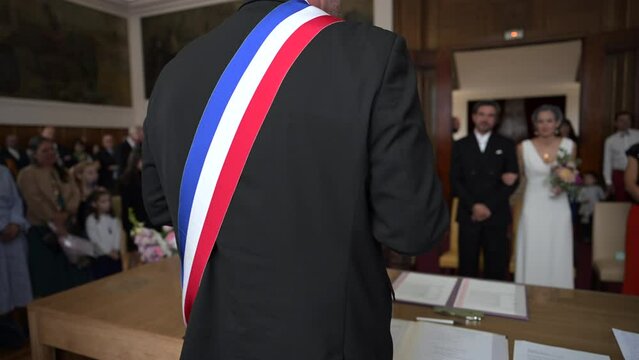 A mayor from behind with a French tricolor scarf making a speech to celebrate a heterosexual marriage in a town hall. Blurred background on the bride and groom. French wedding with two happy lovers.