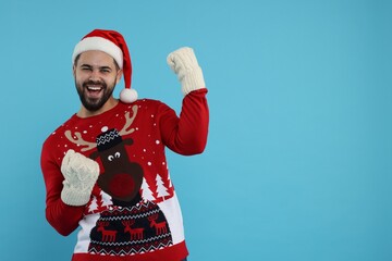 Obraz premium Happy young man in Christmas sweater, Santa hat on and knitted mittens against light blue background. Space for text