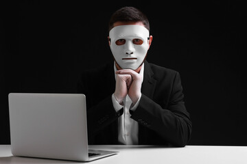 Man in mask and gloves sitting with laptop at white table against black background