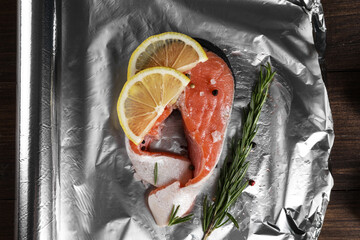 Aluminum foil with raw fish, lemon slices, rosemary and spices on wooden table, flat lay. Baking...