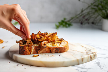 Female hand making a toast with roasted chanterelles and cheese, on marble kitchen countertop. Fried wild mushrooms on toasted slice of bread. - obrazy, fototapety, plakaty
