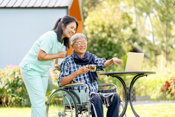 Senior man on wheelchair point to monitor of laptop and talk with Asian nurse who take care him to get good health outdoor.