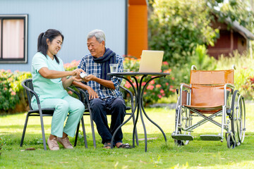 Asian nurse woman give the medicine pill from bottle to senior man and they sit on chair in the garden with wheelchair is set beside.