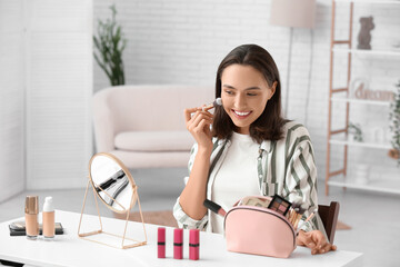 Beautiful young woman with cosmetic bag doing makeup at table in dressing room