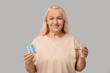 Beautiful mature woman with pills and glass of water on grey background