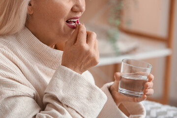 Beautiful mature woman with glass of water taking pill at home