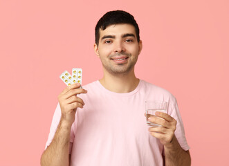 Young man with blisters of vitamin A pills and water on pink background