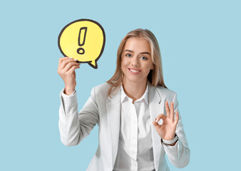 Young businesswoman holding speech bubble with exclamation mark and showing OK gesture on blue background. Business idea concept