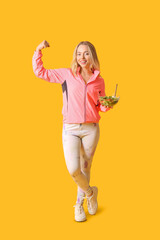 Beautiful sporty woman with bowl of fresh vegetable salad showing muscles on yellow background. Weight loss concept