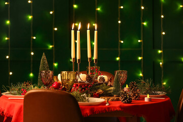 Festive dining table set for Christmas with burning candles and fir cones in room at night