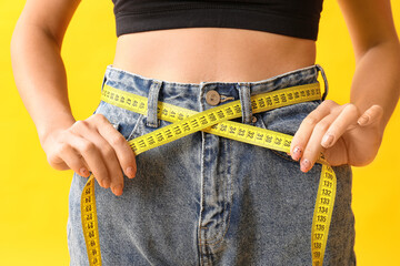 Beautiful young Asian woman with measuring tape on yellow background, closeup. Weight loss concept