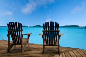 Two weathered Muskoka chairs elegantly poised on a rustic dock, framing a tranquil expanse of...
