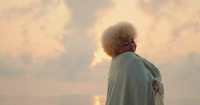 A young African American woman with curly blond hair is wrapped in a blanket and baskin near the ocean at dawn. Cozily wrapped in a warm blanket on the beach. Meet the first rays of the sun.