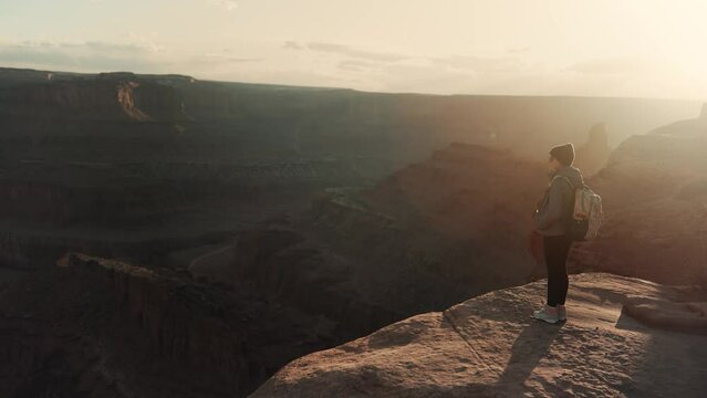 Female hiker walks to the edge of a cliff and admires stunning canyon views during sunset