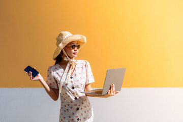 Asian businesswoman, wearing glasses, hat and dress, holding a smartphone and using a laptop for work, standing, relaxing, on vacation near a yellow wall. At a hotel in a beautiful location