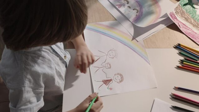 Boy at his desk, using his artistic skills to paint picture of his family with bright rainbow. This drawing depicts happy family, reflecting child's perception of loving and harmonious home, adoption