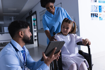 Diverse male and female doctors talking with girl in wheelchair using tablet in hospital corridor