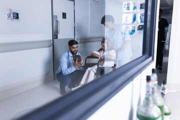 Diverse male and female doctors talking with girl in wheelchair using tablet in hospital corridor