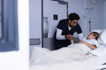 Obraz premium Diverse male doctor talking with girl lying in bed in hospital corridor