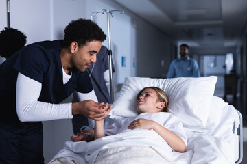 Diverse male doctor talking with girl lying in bed in hospital corridor