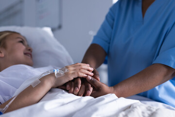Fototapeta na wymiar Midsection of diverse female doctor holding hands of girl lying in bed in hospital corridor