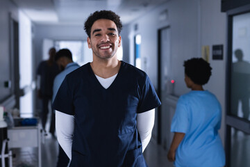 Portrait of happy biracial male doctor standing in hospital corridor - obrazy, fototapety, plakaty