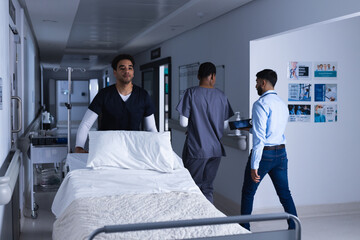 Biracial male doctor walking with bed in hospital corridor - Powered by Adobe