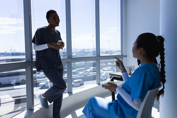 Diverse male and female doctors talking, drinking coffee and eating lunch in sunny hospital