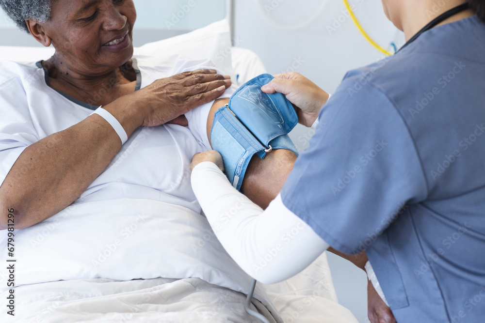 Wall mural Diverse female doctor testing blood pressure of senior female patient in hospital room