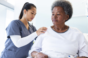 Diverse female doctor testing senior female patient using stethoscope in hospital room