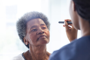 African american female doctor testing eyes of senior female patient in hospital room - obrazy, fototapety, plakaty