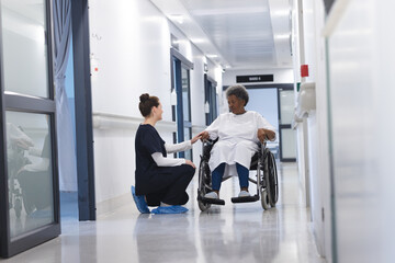 Diverse female doctor talking with senior female patient in wheelchair in hospital corridor