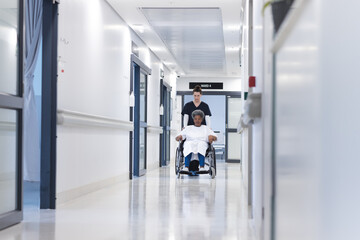 Obraz premium Diverse female doctor walking with senior female patient in wheelchair in hospital corridor