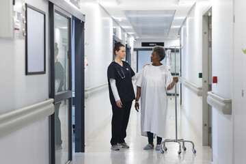 Diverse female doctor talking with senior female patient with drip in hospital corridor