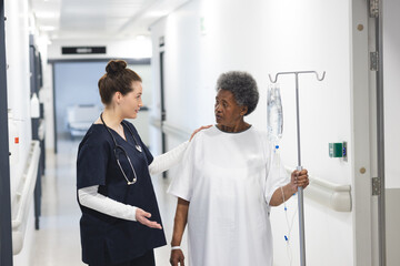 Diverse female doctor talking with senior female patient with drip in hospital corridor