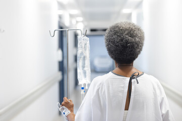 Back of african american senior female patient with drip walking in hospital corridor