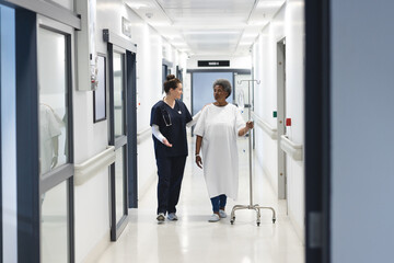 Happy diverse female doctor talking with senior female patient with drip in hospital corridor