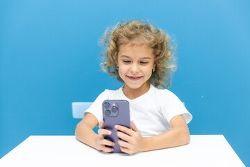 Kid and gadget. Happy laughing little child girl sitting at desk holding mobile phone in hands, making call to mother, chatting with friend, watching funny cartoon, posting selfie in social network