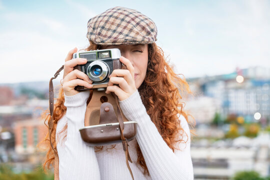 Portrait of an excited and happy young lady with red curly hair exploring city  and taking pictures on an old film camera