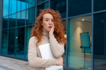 unhappy redhead woman holding laptop and in shock from new in city center