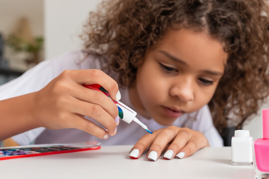 little girl paints her nails. a girl with short curly hair in a white T-shirt sits at a table and paints her nails with white polish