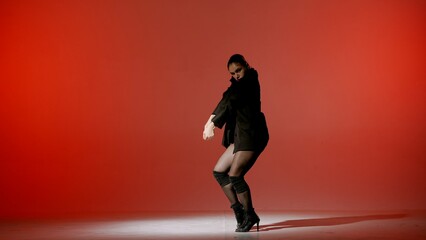 On a red background. Young woman, showing dance moves towards high heels. A spotlight beam shines on her and gives her a shadow, the edges of the background are darkened. She is sexy, rhythmic