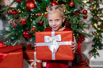 Cute girl with a gift in her hands sits near the Christmas tree.