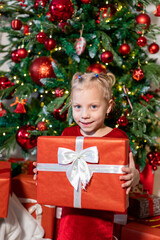 Cute girl with a gift in her hands sits near the Christmas tree.