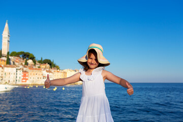Fototapeta na wymiar Funny little girl posing on embankment sunny summer day. Rovinj town in background. Travel and adventure concept.