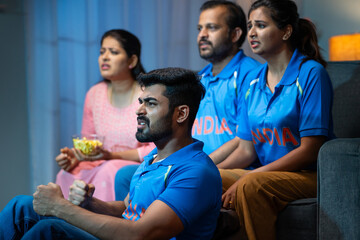 Tensed Senior Middle aged parents with siblings watching live Cricket sports match with Indian Tshirts at home on sofa - concept of entertainment, Nail biting and intense expression.